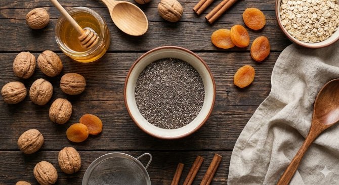 Top view of healthy baking ingredients including chia seeds, walnuts, honey, and dried apricots arranged on a dark wooden table